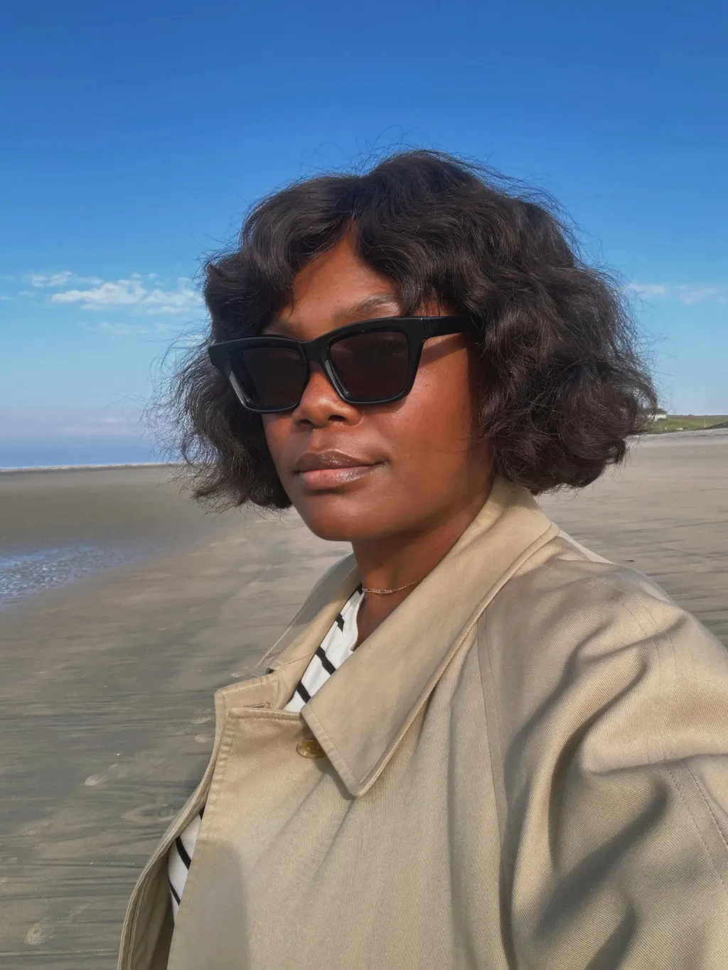 Frau mit lockigem Bob am Strand, dazu trägt sie eine Sonnenbrille
