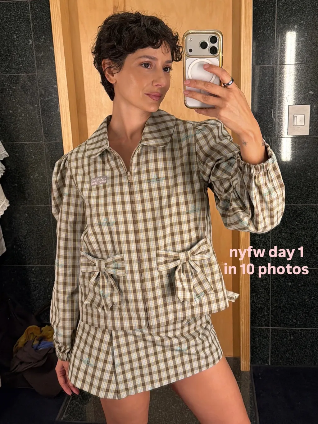 Frau mit lockigem Pixie-Schnitt in kariertem Hemd und Rock, die ein Selfie im Spiegel macht